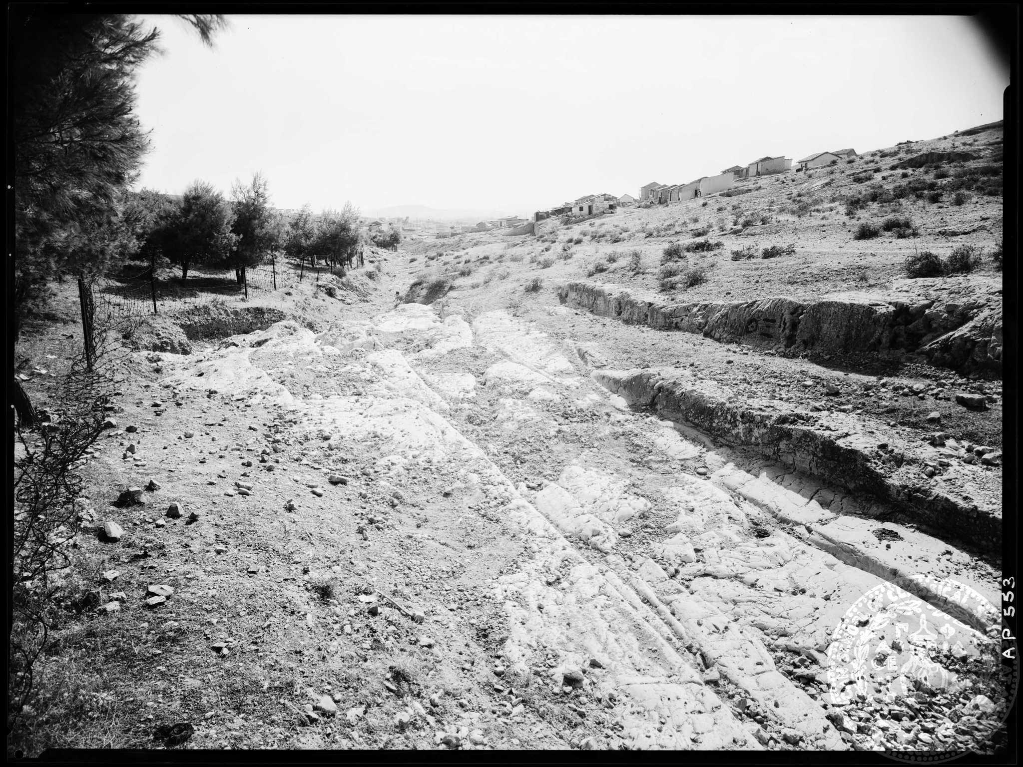 Το βλέμμα ανηφορίζει στα σπιτάκια του Ασυρμάτου, πέρα από τον δρόμο με τις αρχαίες αρματροχιές (Αμερικανική Σχολή Κλασικών Σπουδών στην Αθήνα, Αρχαιολογικό Φωτογραφικό Αρχείο, AP 0553).